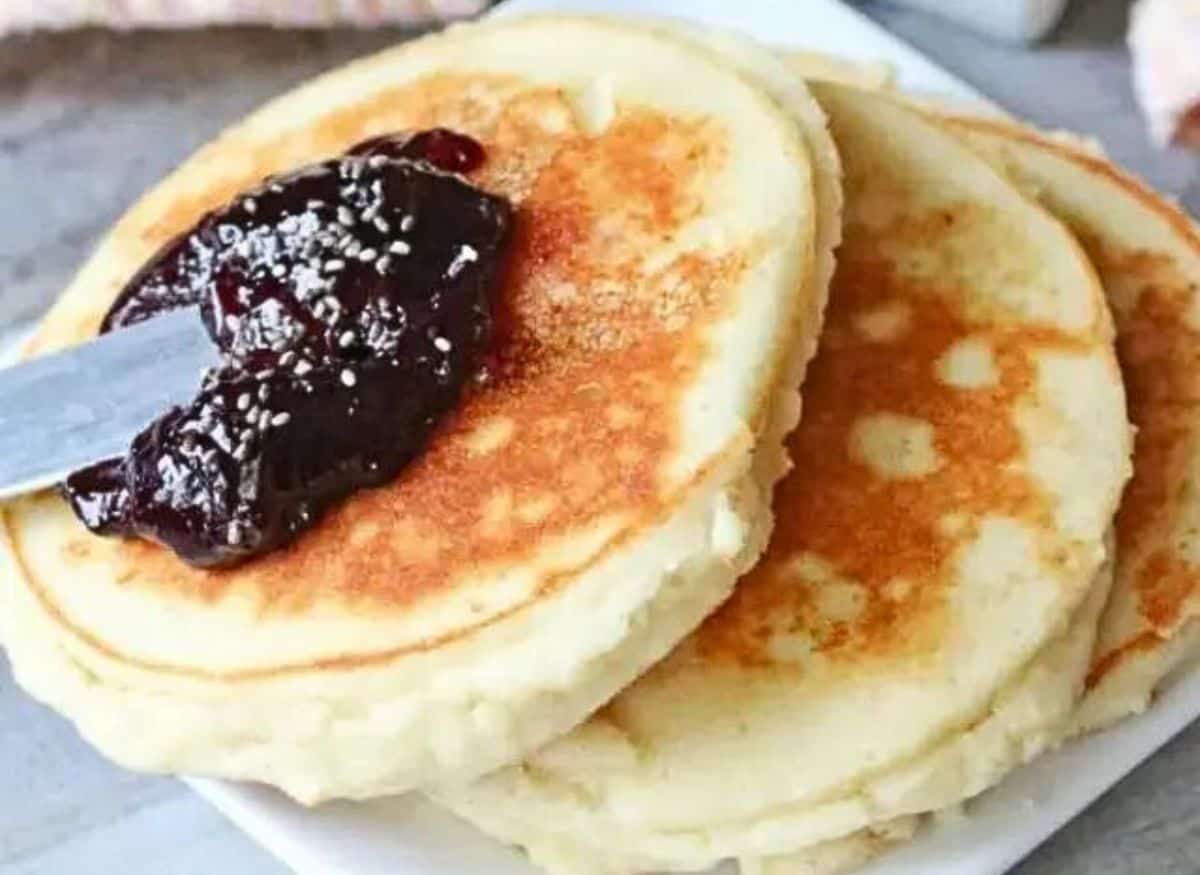 Three yogurt pancakes on a white plate topped with jam. Syrup in a white container behind it and a light orange napkin