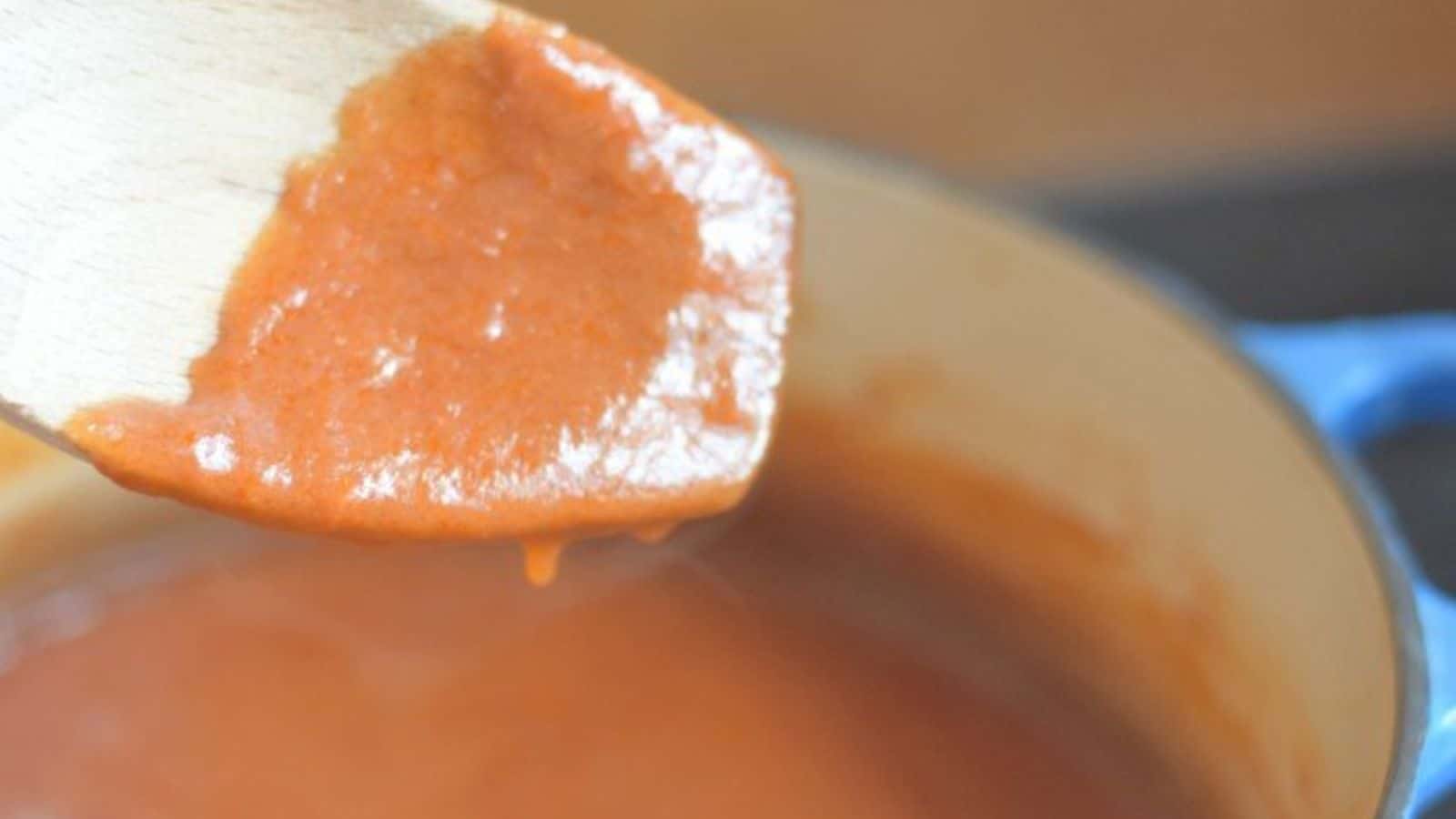 Image shows A wooden spoon is dripping red enchilada sauce into the pot containing the rest.