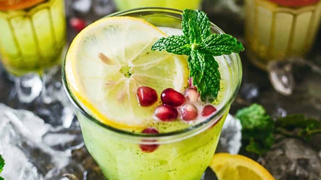 A glass of bright green mojito garnished with a lemon slice, pomegranate seeds, and mint leaves. Several decorative glasses are in the background. Lemon wedges and ice cubes surround the glass on the table.