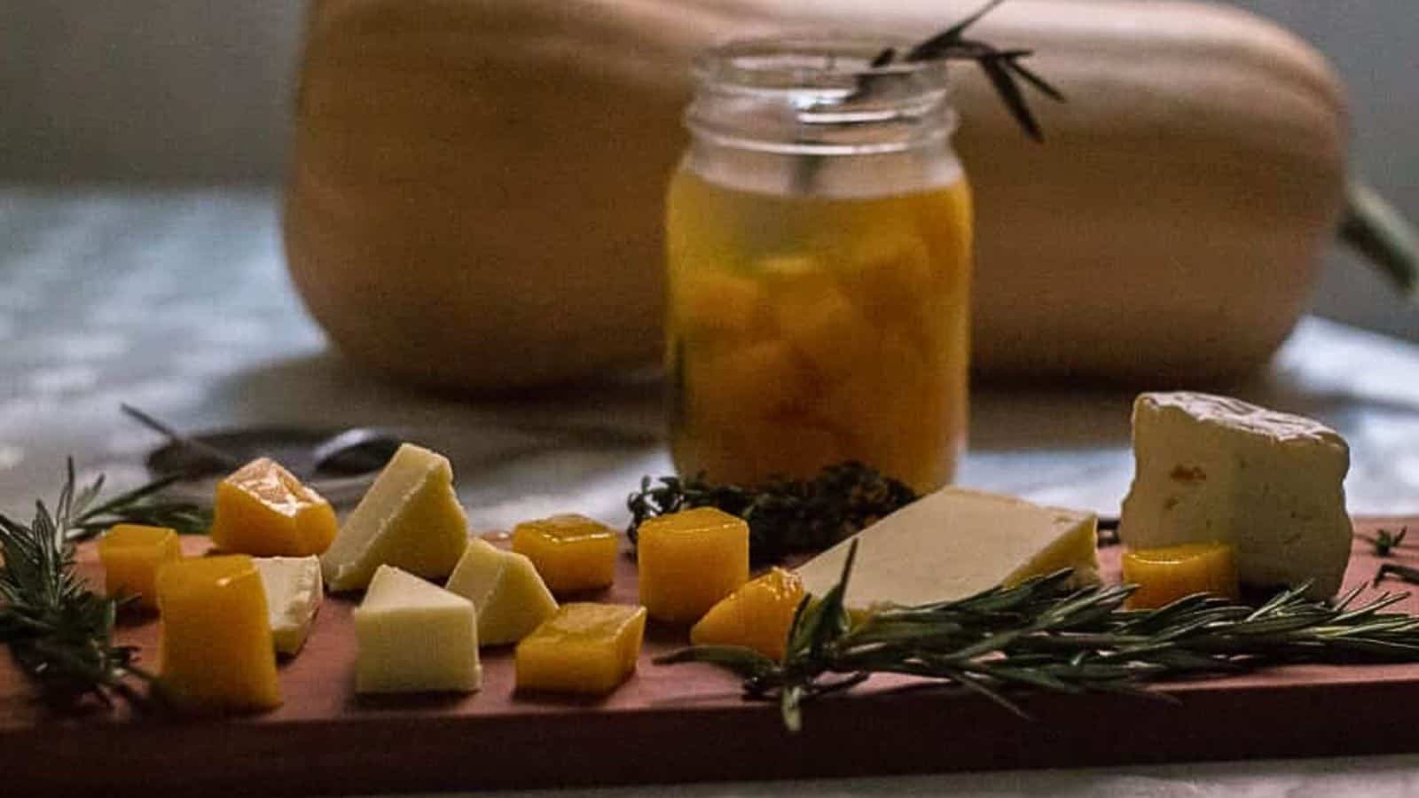 A wooden cutting board with cheese, herbs and a pumpkin.