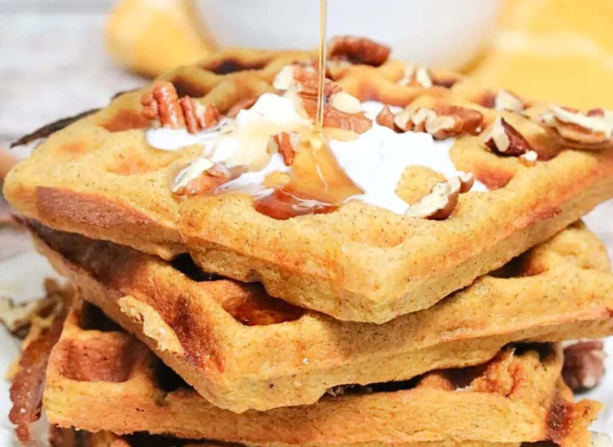 three gluten-free pumpkin waffles stacked on a white plate with coconut cream, maple syrup and pecans on top. A waffle maker beside the plate and a bowl of pecans and syrup behind it