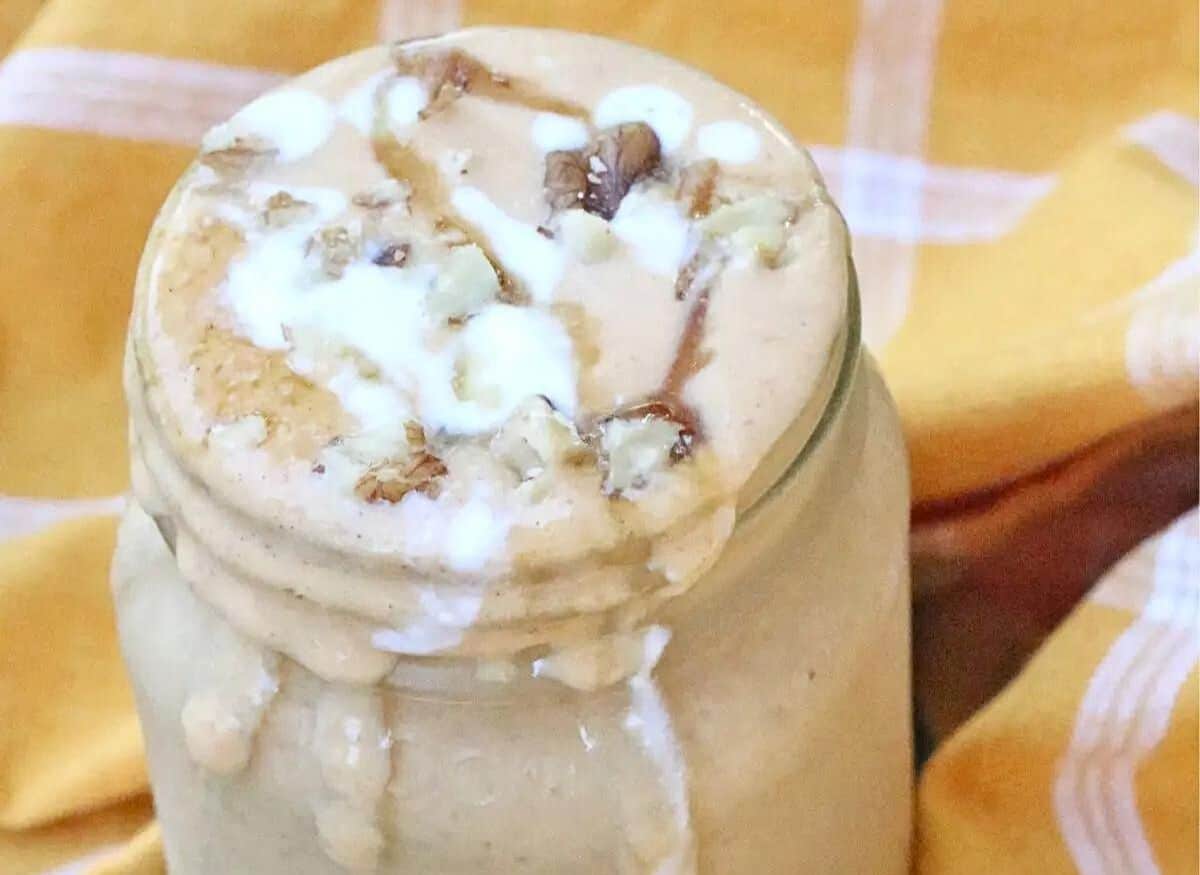 Mason jar with light orange pumpkin smoothie tipped with coconut whip cream, nuts and a blue straw spilling down the side.