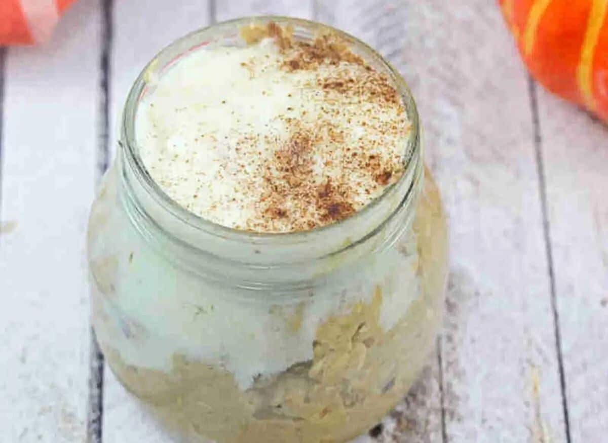 Mason jar with pumpkin spice overnight oats with cream cheese frosting on top and an orange linen and decorative pumpkins behind it.