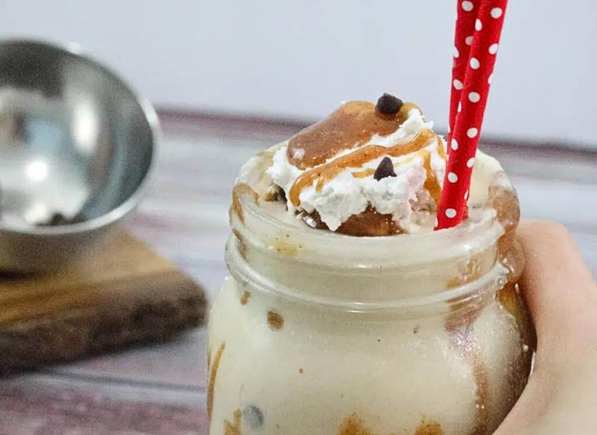 Mason jar with caramel protein shake topped with coconut whipped cream, caramel sauce and chocolate chips two red straws in the smoothie and chocolate chips on a cutting board behind it.