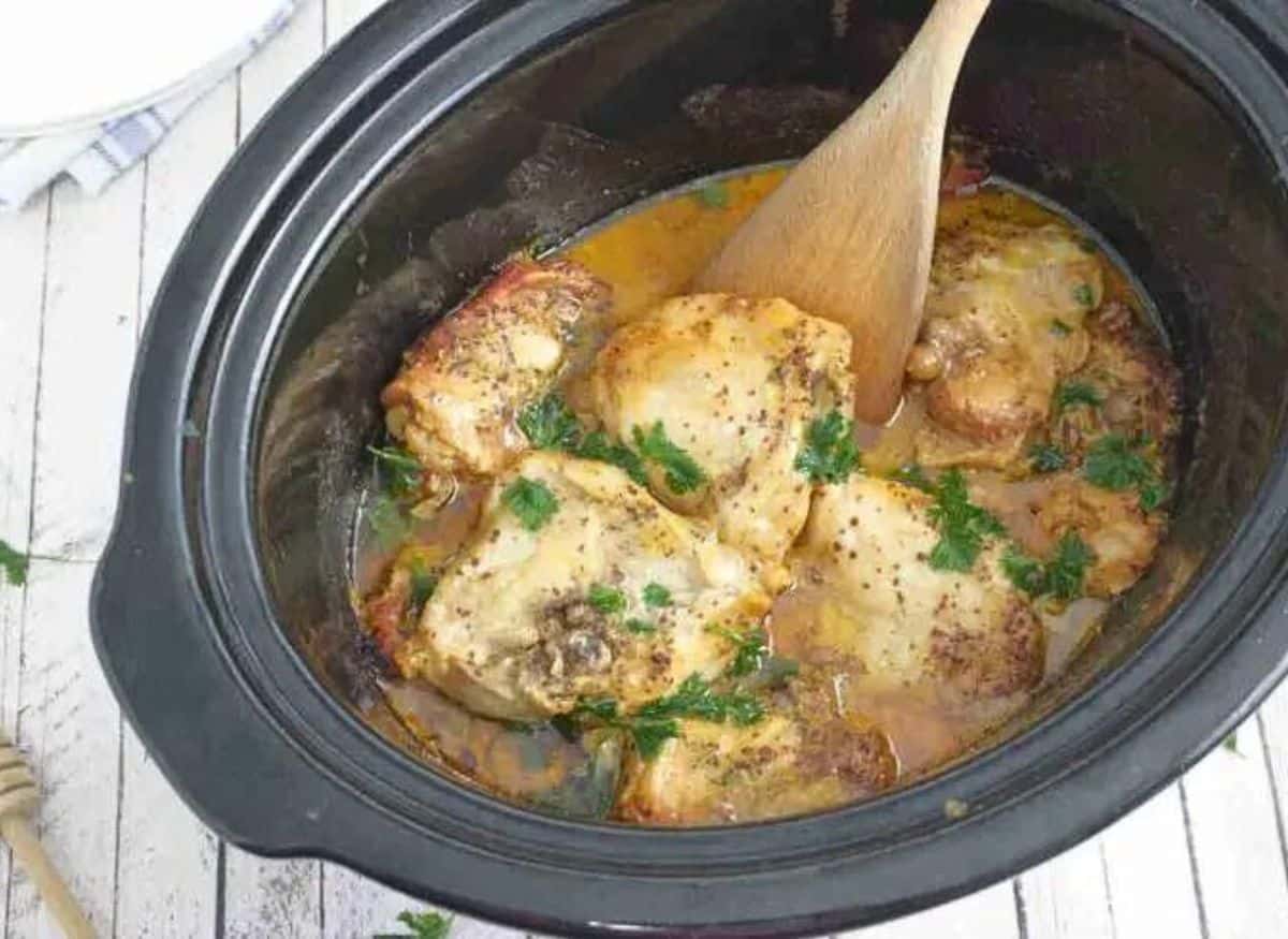 Black slow cooker with honey mustard chicken thighs topped with parsley and a wooden spoon in it.