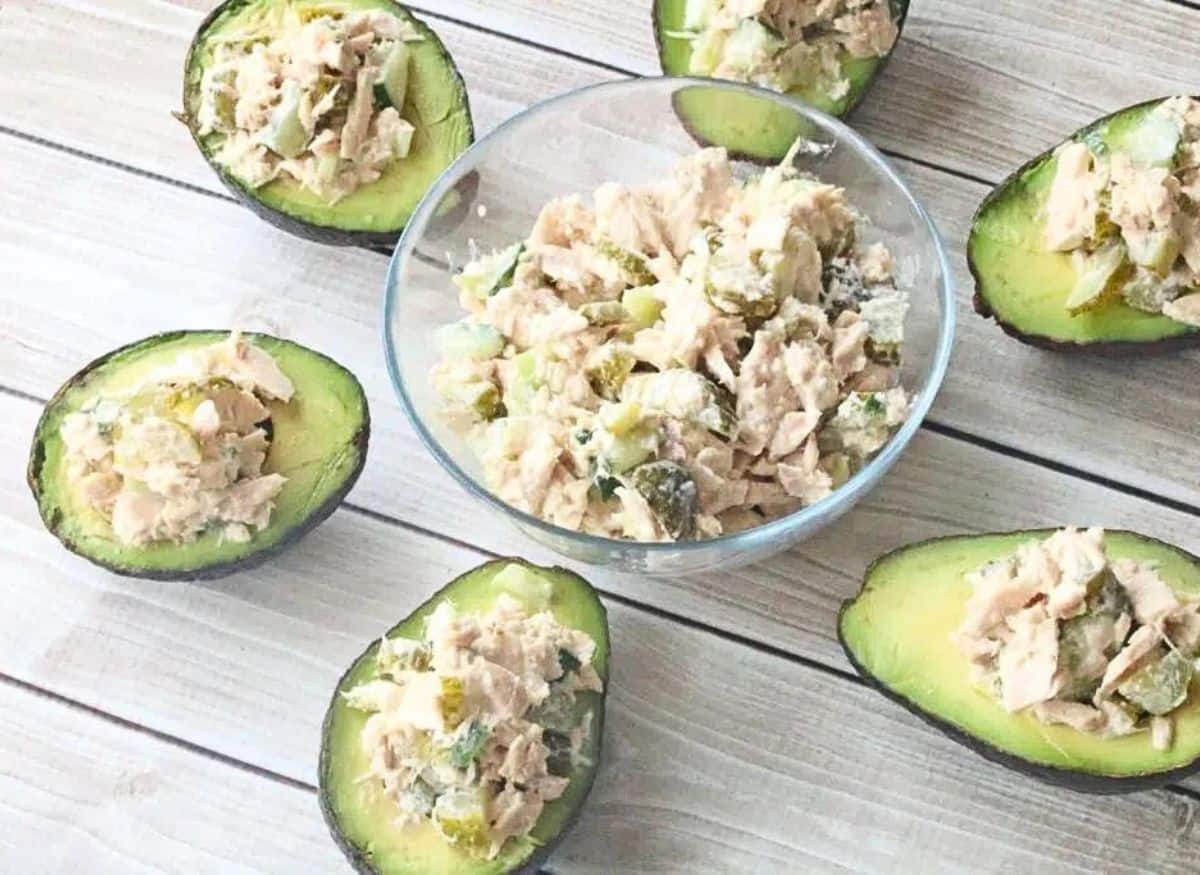 A glass bowl of tuna salad with pickles and multiple avocados stuffed with the salad around it.