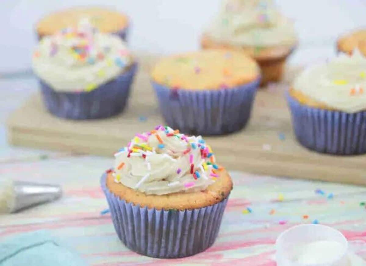 Vanilla protein cupcake with dairy-free frosting and sprinkles in a blue cupcake liner with more cupcakes behind it on a cutting board.