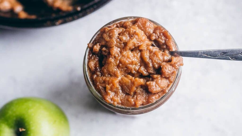 Apple butter in a jar with a spoon next to an apple.