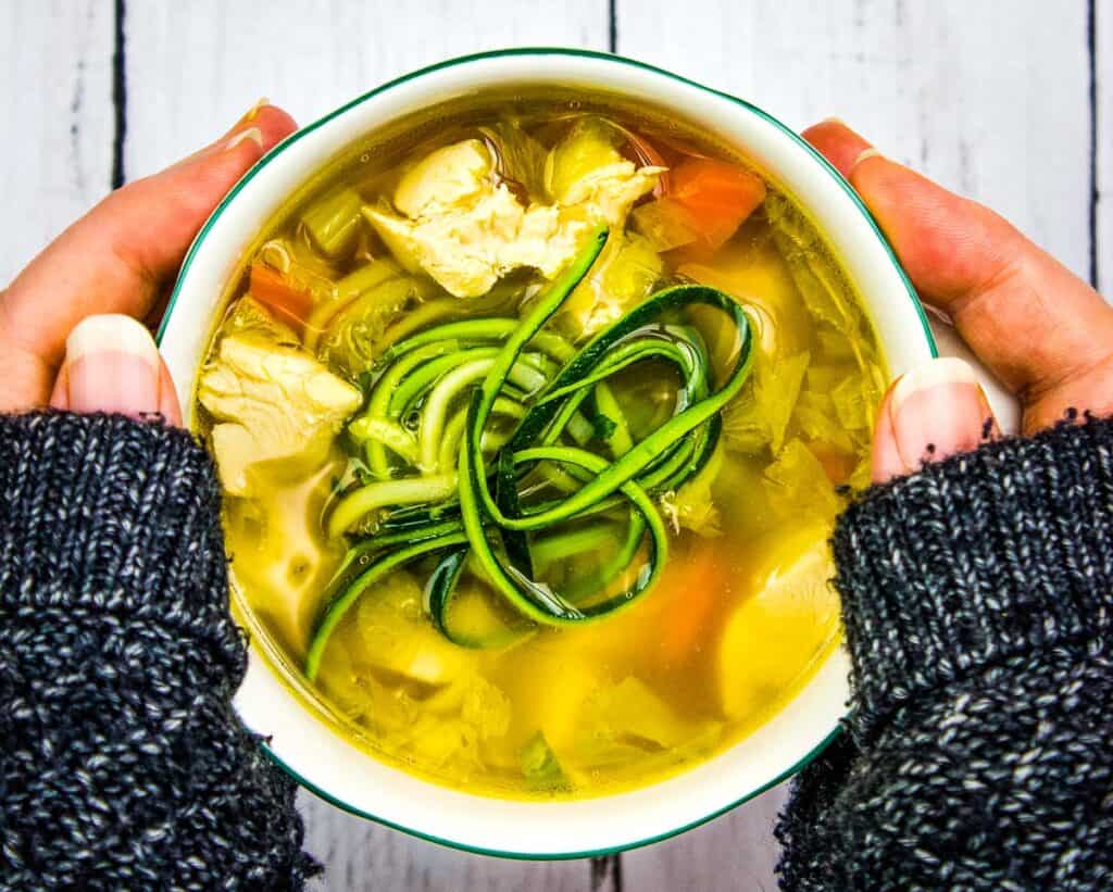 A person holding a bowl of chicken and zucchini soup.
