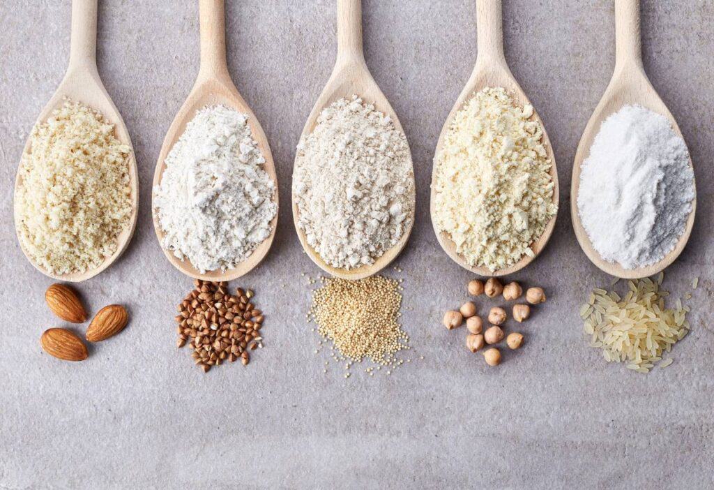 Different types of gluten-free flour in wooden spoons on a gray background.