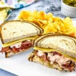 A Pastrami Reuben sandwich on a plate with chips and pickles.