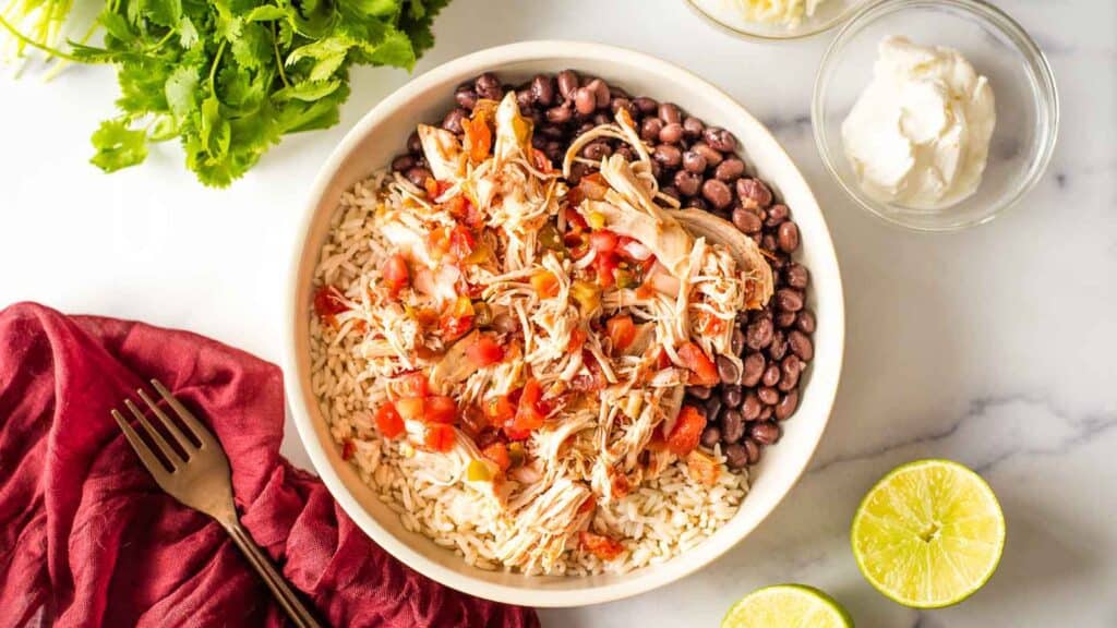 A bowl of slow cooker salsa chicken, mexican rice, black beans and limes.