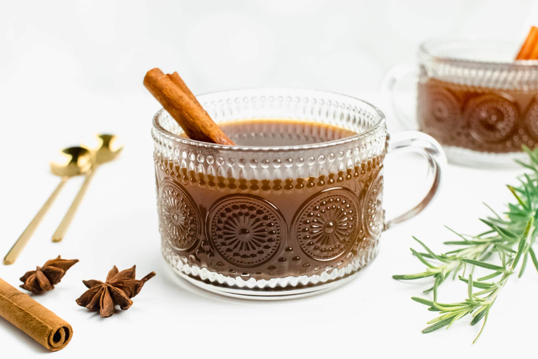A glass mug holds a warm cider drink surrounded by whole spices and rosemary sprigs.