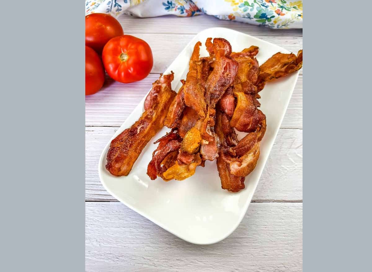Bacon strips on a white plate next to tomatoes.