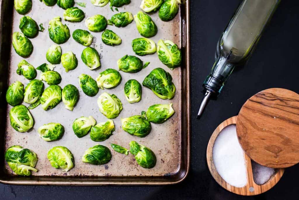 Brussels sprouts on a sheet pan next to a bottle of oil and a bowl of salt.
