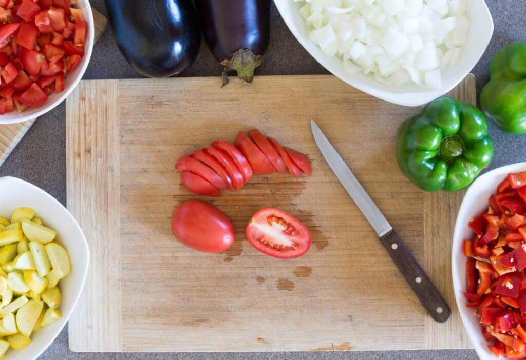 Sliced tomatoes on a cutting board with a knife surrounded by chopped vegetables.