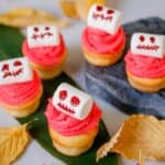 A group of Halloween cupcakes with red frosting and skeleton marshmallows on a table decorated with leaves.