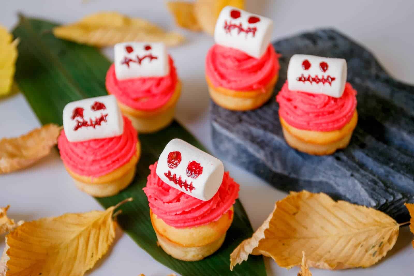 A group of Halloween cupcakes with red frosting and skeleton marshmallows on a table decorated with leaves.