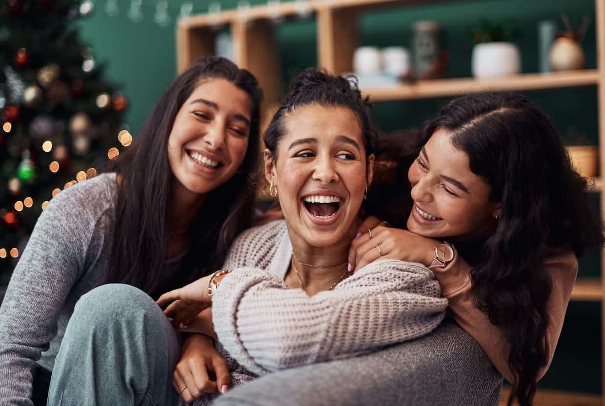 Three women laughing while sitting on a couch in front of a christmas tree.