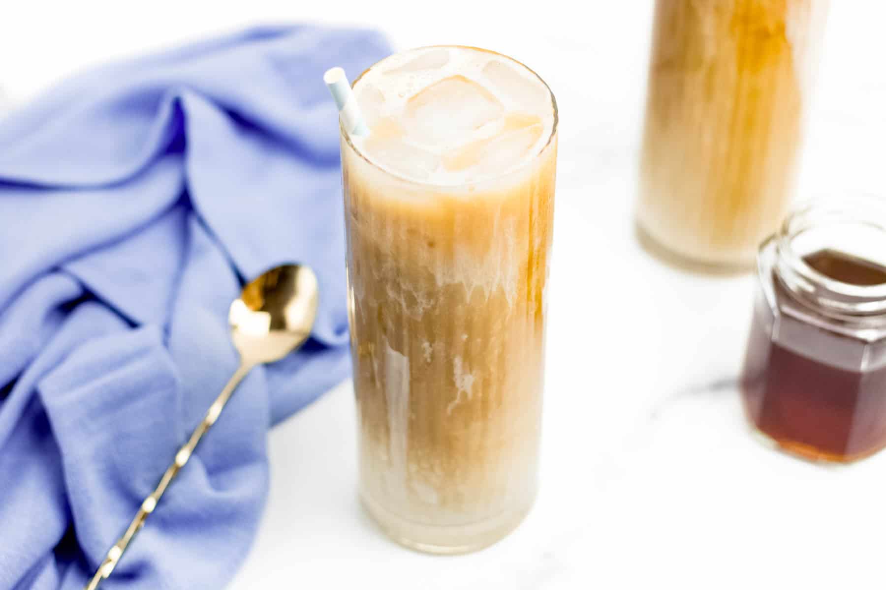 Two Iced Brown Sugar Oat Milk Shaken Espresso coffee drinks with a gold spoon and blue towel and a jar of brown sugar syrup.
