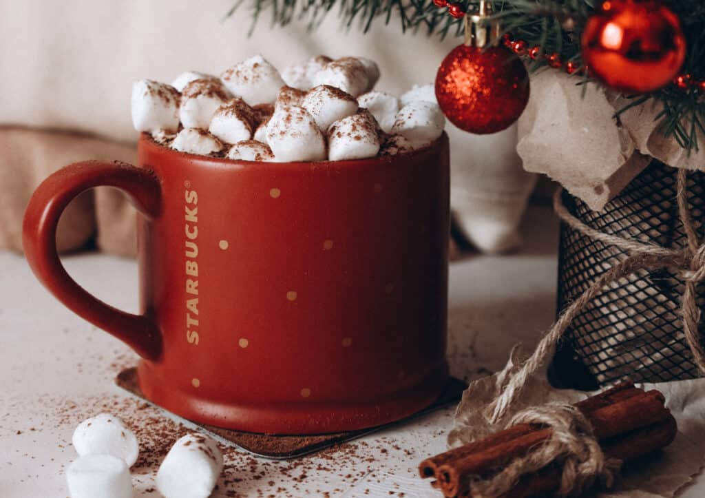 A starbucks mug with marshmallows and cinnamon near a christmas tree.