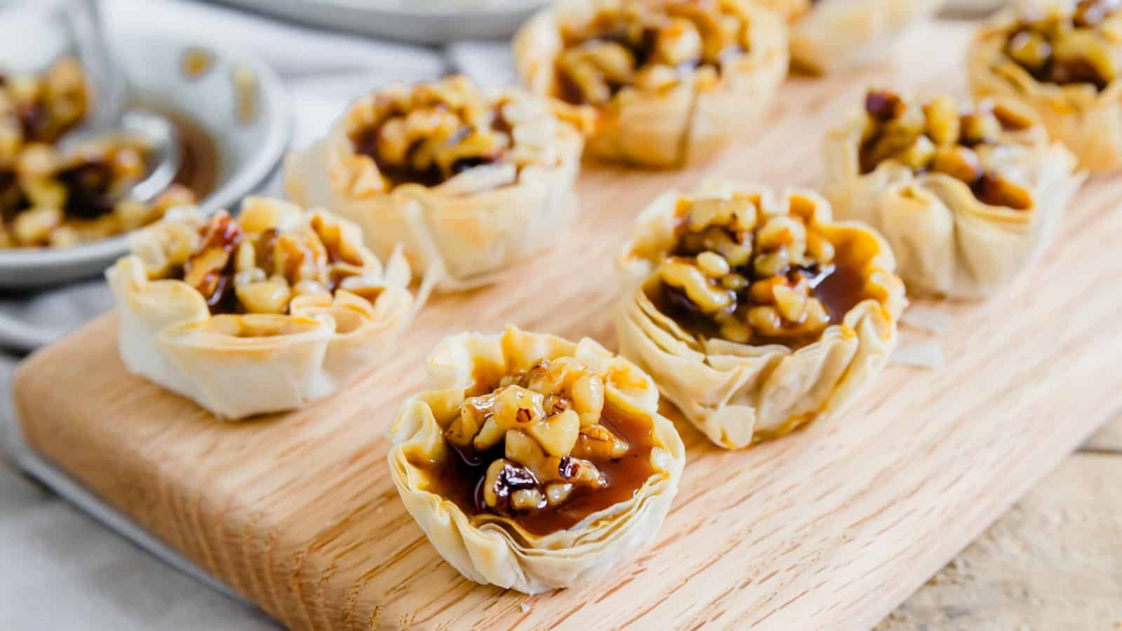 A wooden cutting board with a tray of mini pumpkin pie bites topped with maple walnuts.