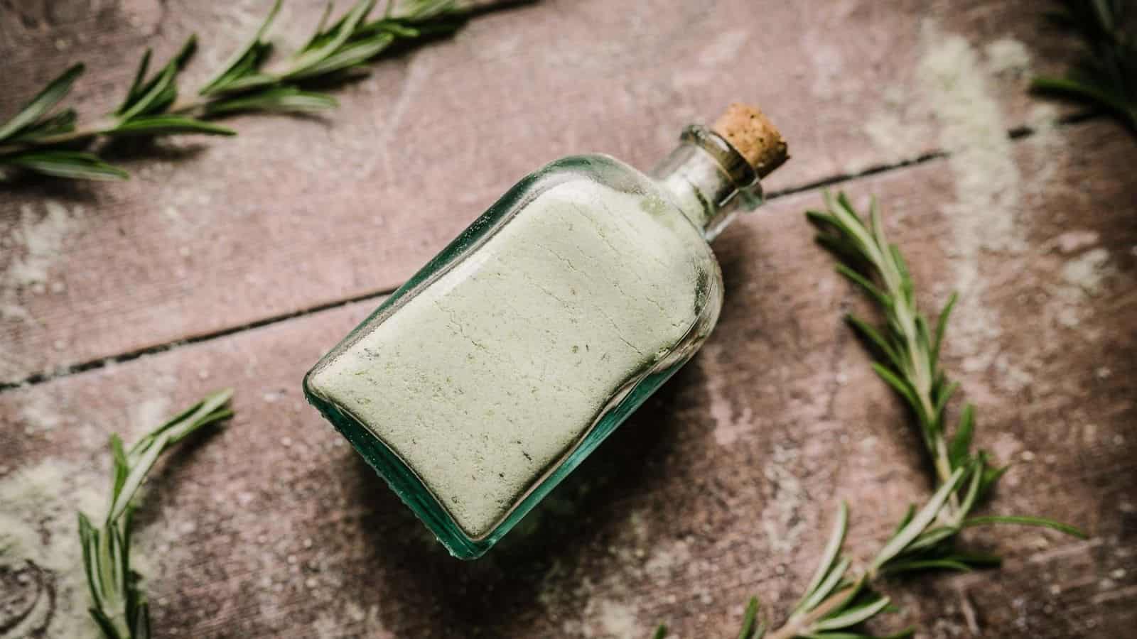 A bottle of green salt with rosemary sprigs on a wooden table.