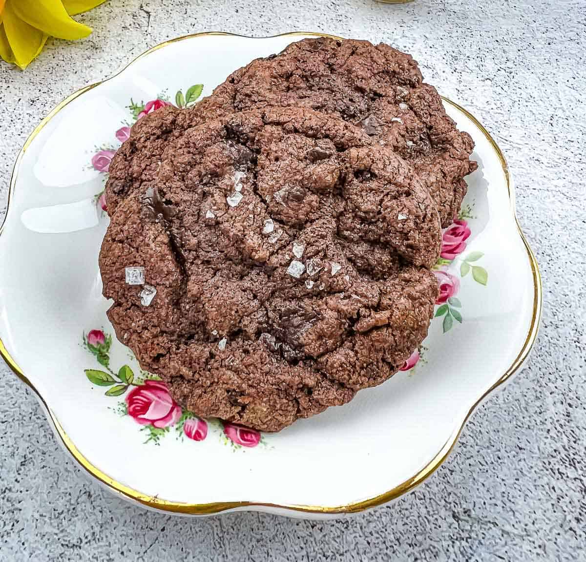 Two Salted Dark Chocolate cookies on a plate next to flowers.