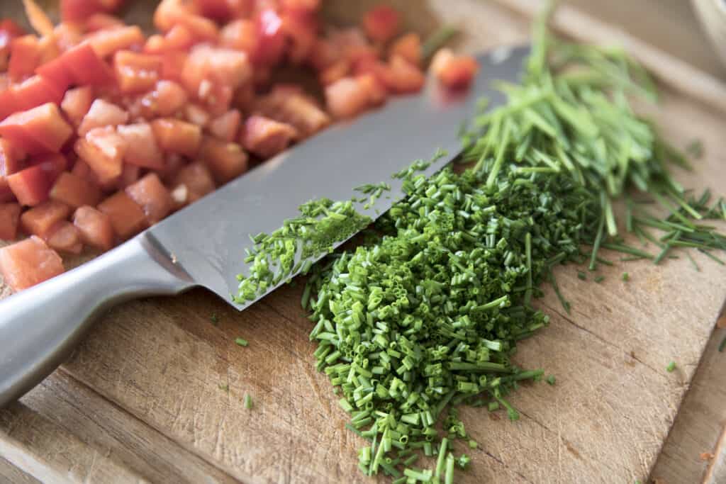 A knife is slicing tomatoes and chives on a cutting board.