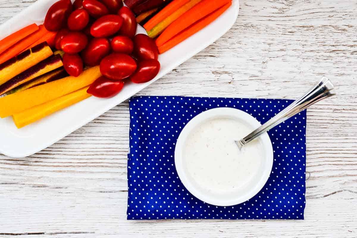 A bowl of creamy garlic dressing with carrots and tomatoes on a blue cloth.
