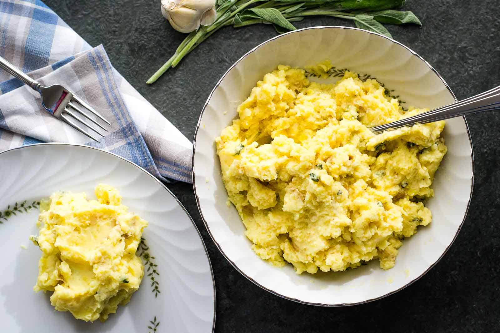 A bowl of brown butter garlic mashed potatoes on a table, next to a serving on a plate.