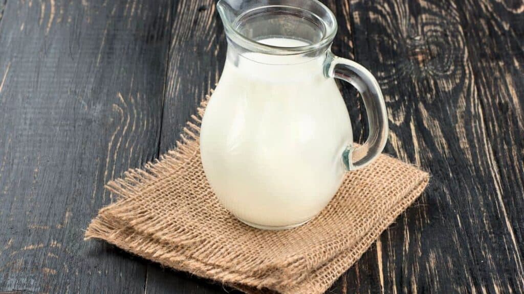 A glass pitcher filled with milk, perfect for experimenting with evaporated milk substitutes, sits on a piece of burlap fabric atop a dark wooden surface.