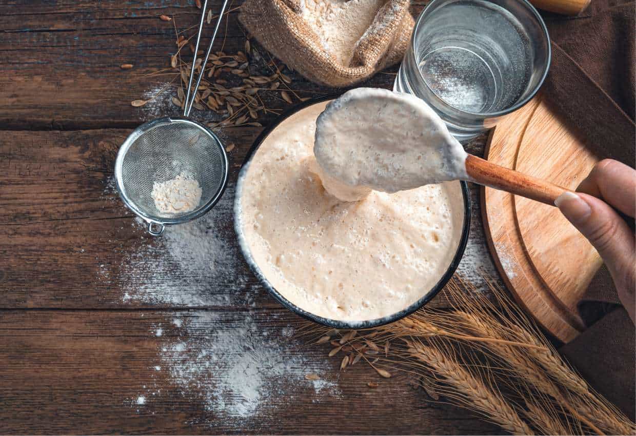 A person scooping out sourdough starter discard from a bowl on a wooden table.