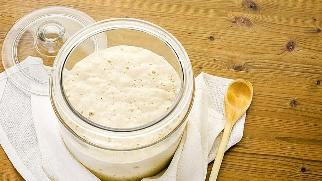 A glass jar filled with bubbly sourdough starter sits on a white cloth next to a wooden spoon on a wooden table, ready to inspire your next batch of sourdough discard recipes.