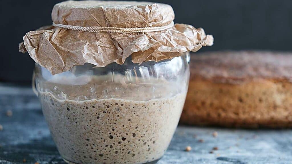 A glass jar of sourdough starter covered with brown paper and tied with string sits on a gray surface, accompanied by a loaf of bread in the background&mdash;perfect inspiration for exploring new sourdough discard recipes.