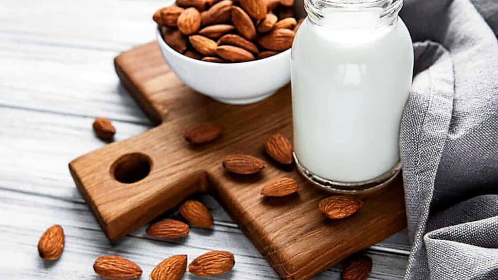 A glass bottle of almond milk, often used as one of the popular evaporated milk substitutes, sits on a wooden board next to a bowl of whole almonds, with scattered almonds and a gray cloth nearby.