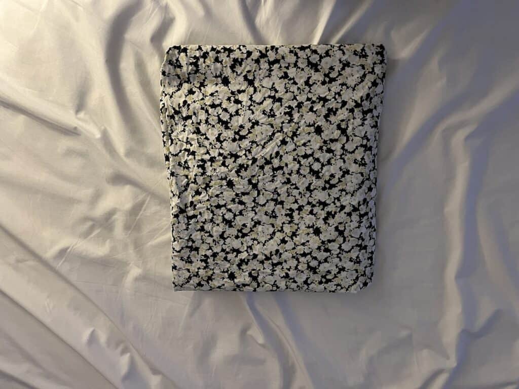 A black and white floral pillow on a white bed with a folded fitted sheet.