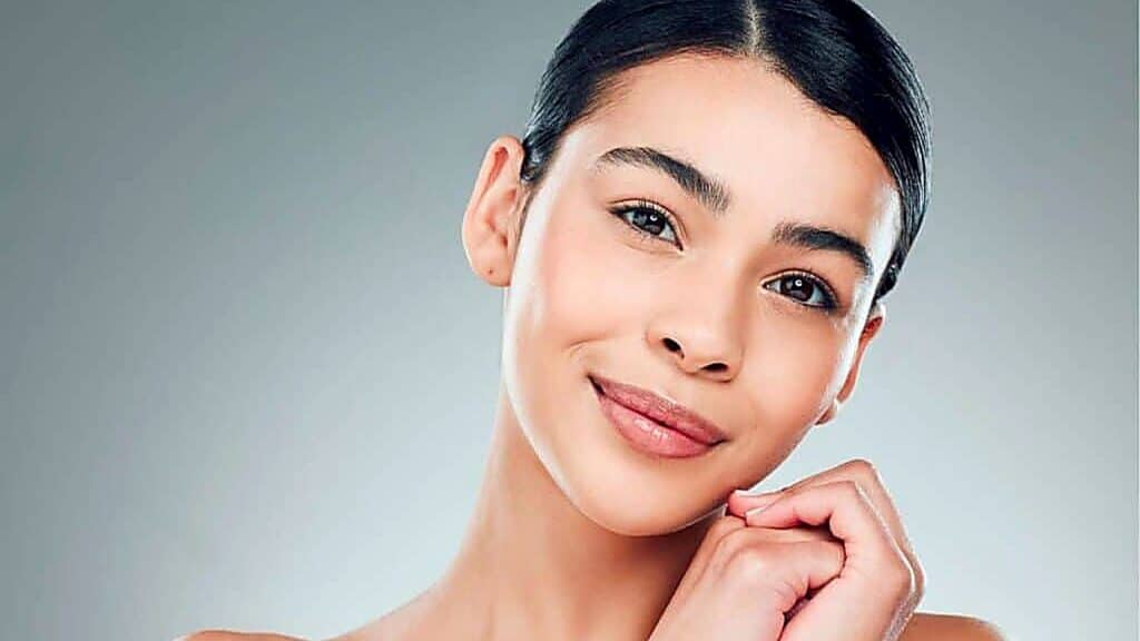 A young woman with clear skin and dark hair pulled back smiles softly, posing against a plain, light background&mdash;her radiant complexion reminiscent of the benefits often associated with enjoying foods high in collagen.