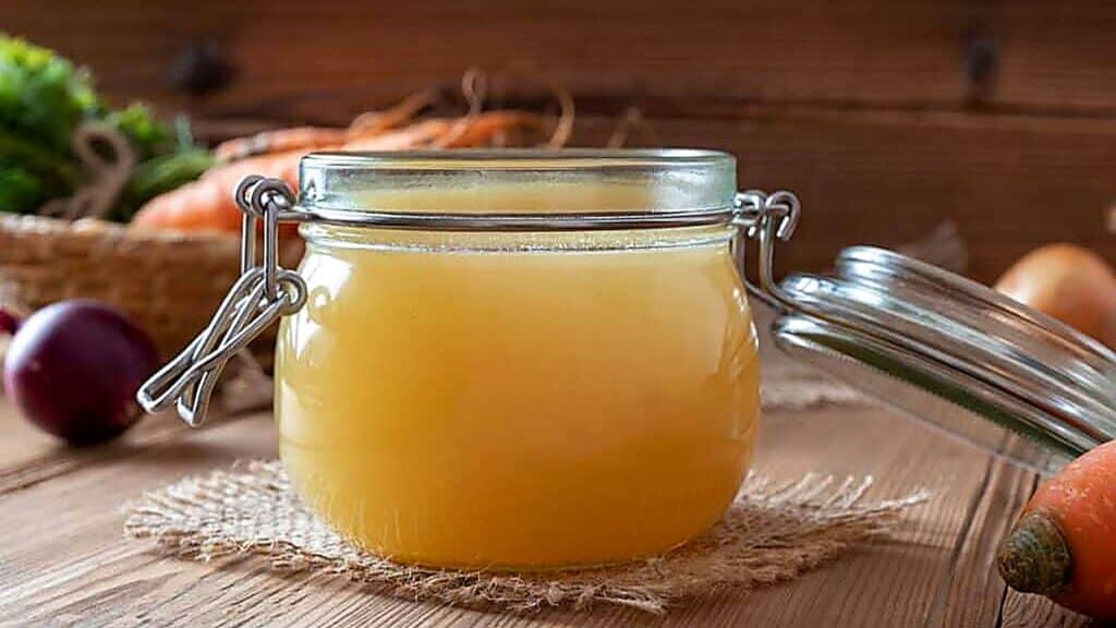 A glass jar filled with light yellow broth, known for being one of the foods high in collagen, sits on a wooden table with vegetables and a basket in the background.