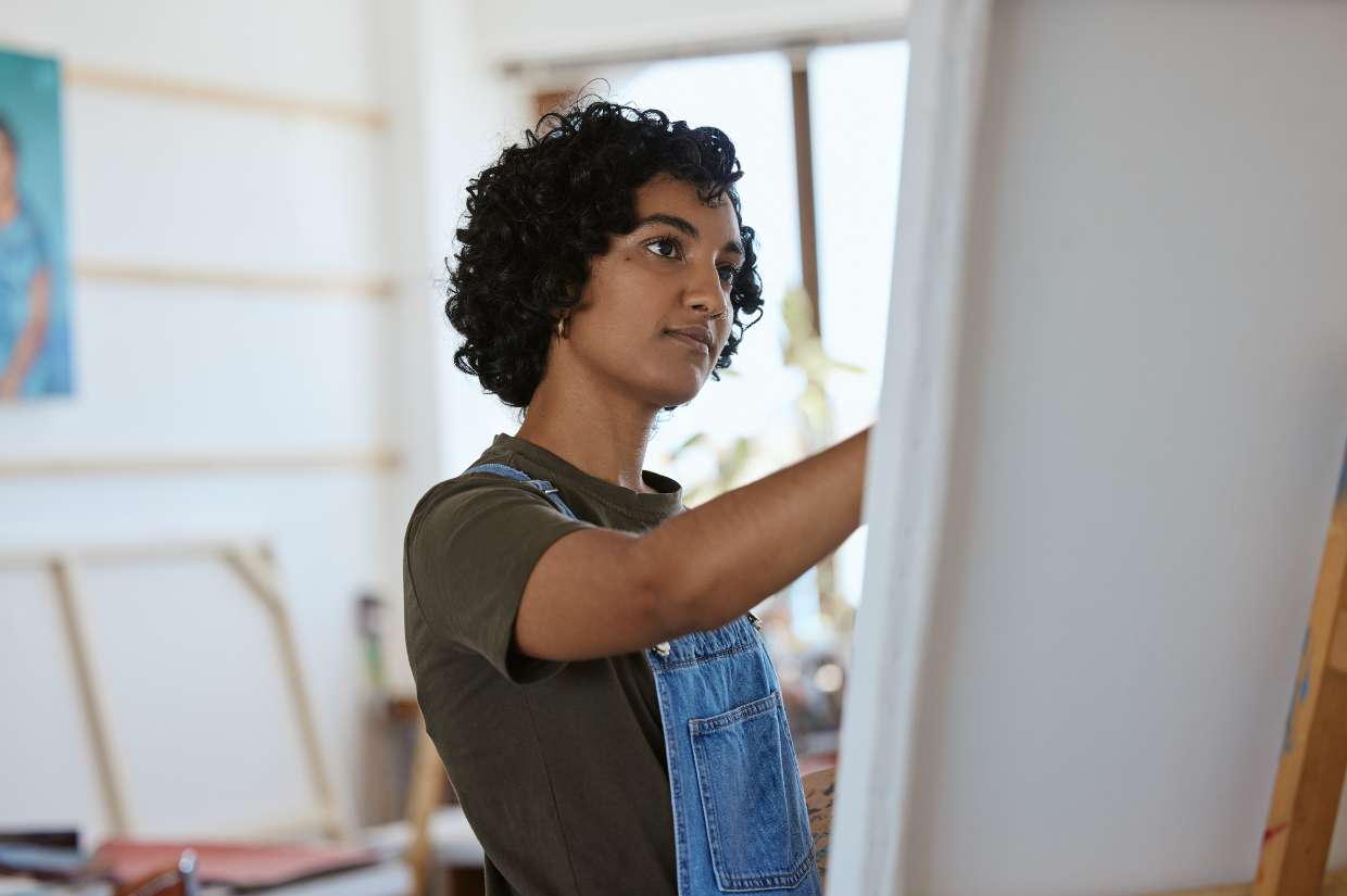 A woman in overalls is engaged in the wellness activity of painting on an easel.