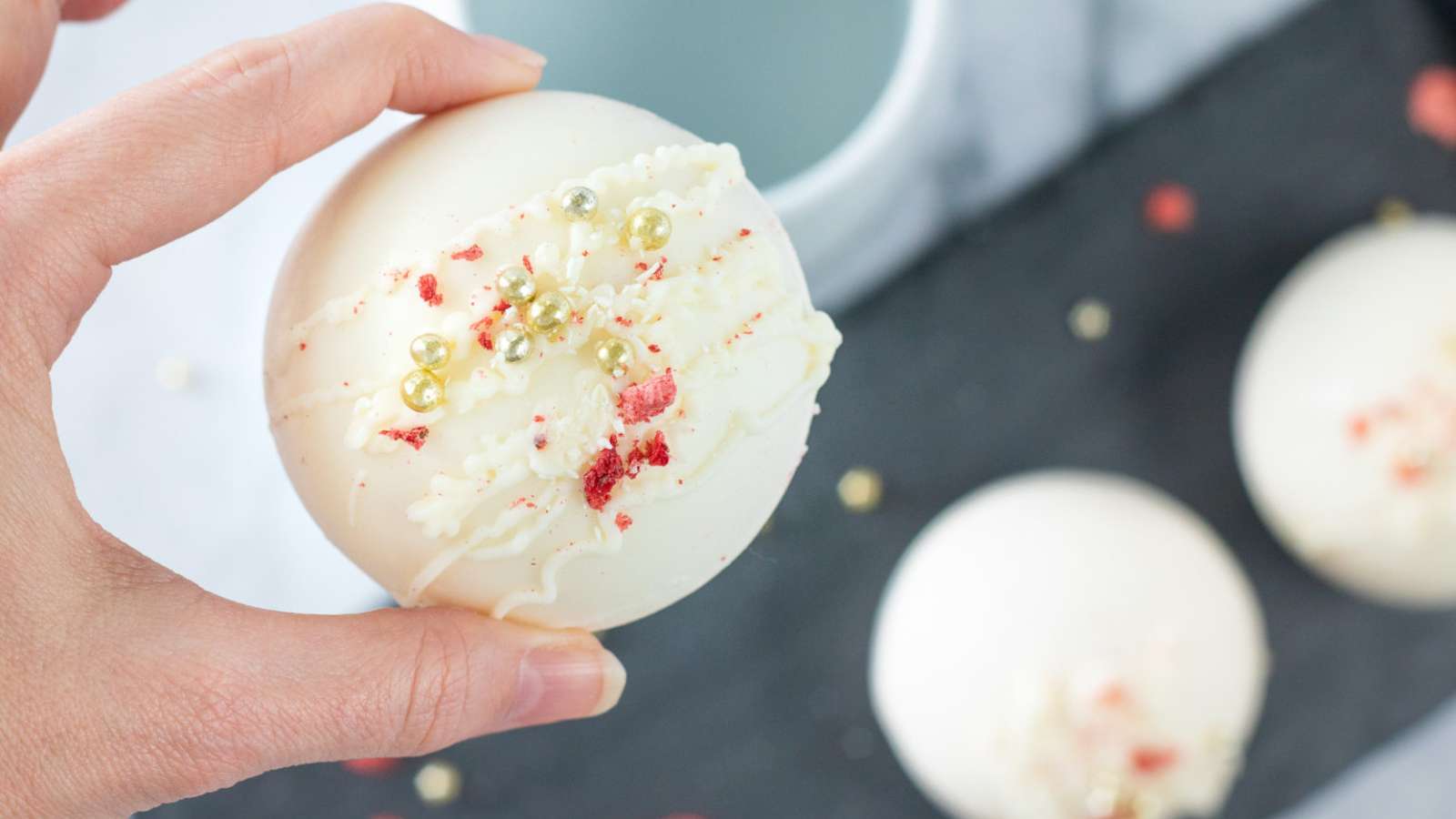 A person holding a Strawberry White Chocolate Cocoa Bomb with sprinkles on it.