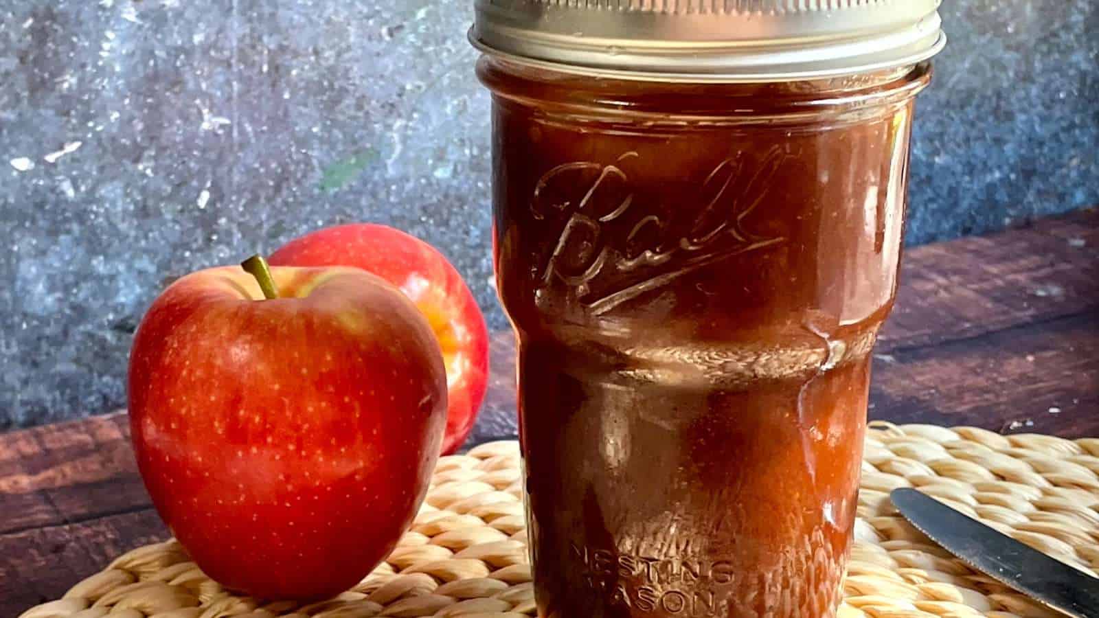 A mason jar of apple butterwith apples and a knife next to it.