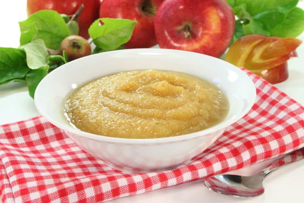 A bowl of applesauce with a spoon and apples.