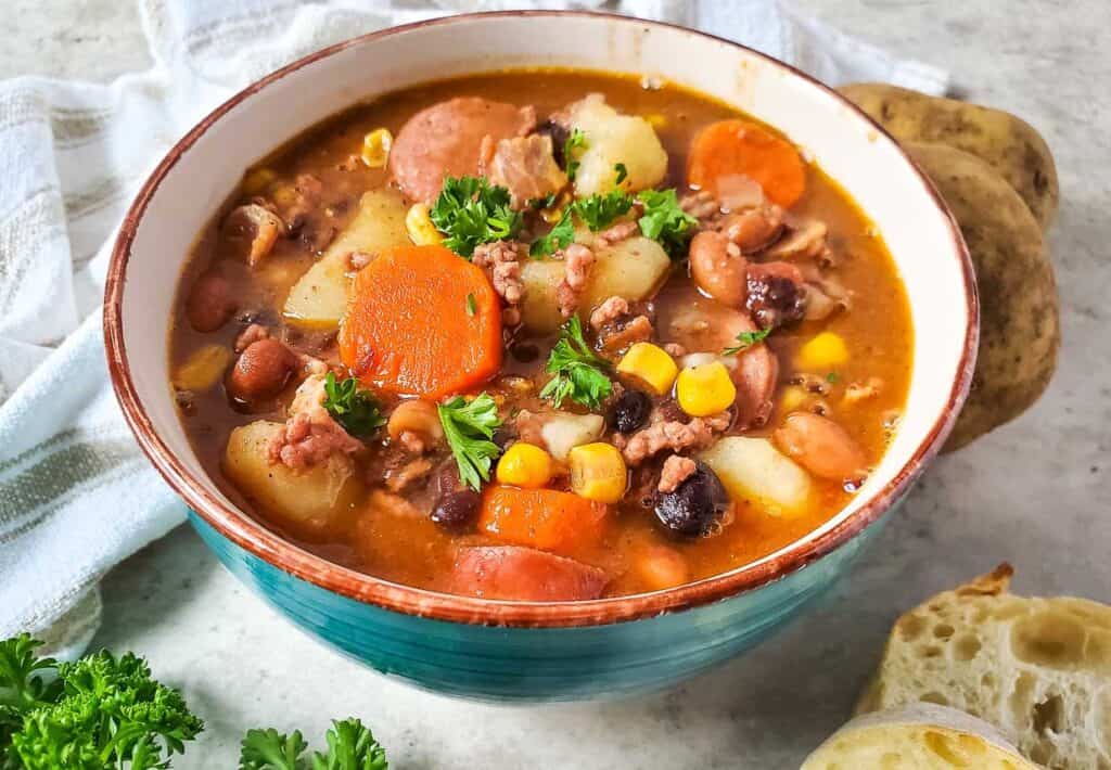 A bowl of soup with meat, potatoes, beans and corn.