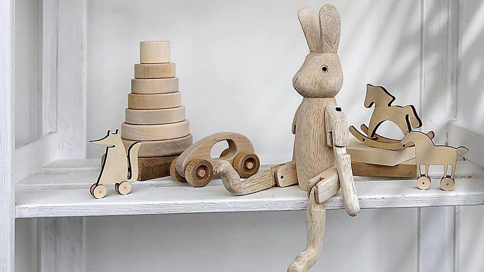 Wooden eco-friendly toys displayed on white shelves, including a stacking tower, a bunny figure, a car, a rocking horse, a cow, and a dog on wheels.