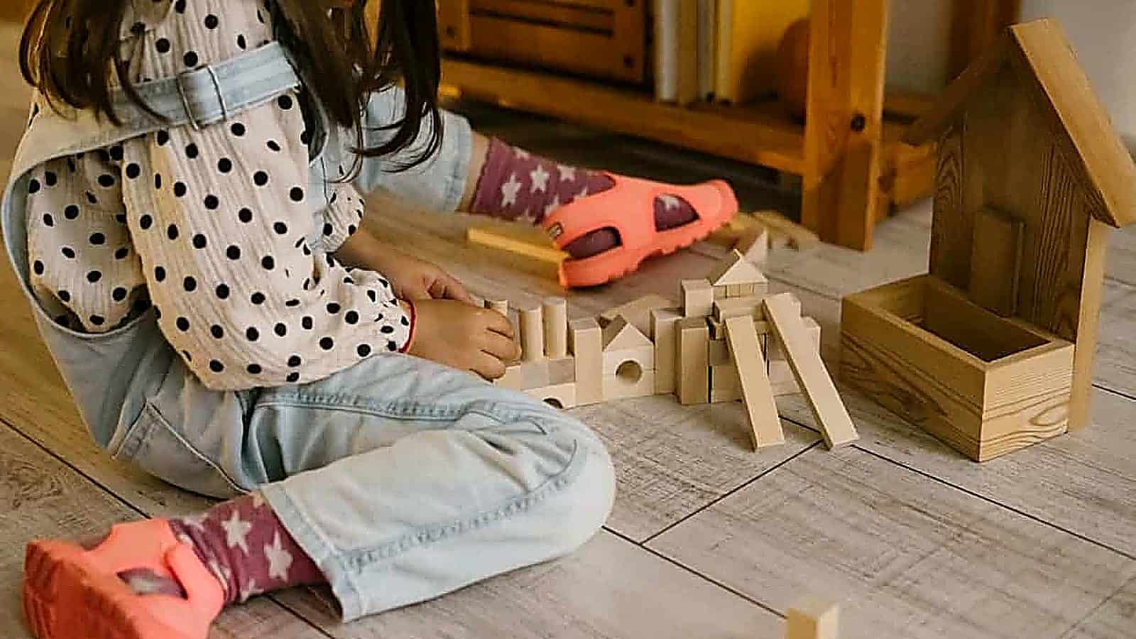 A child in pink shoes and a polka dot shirt sits on the floor, playing with eco-friendly wooden building blocks next to a wooden box and house-shaped structure.