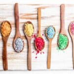 Colorful sprinkles in wooden spoons on a cutting board.