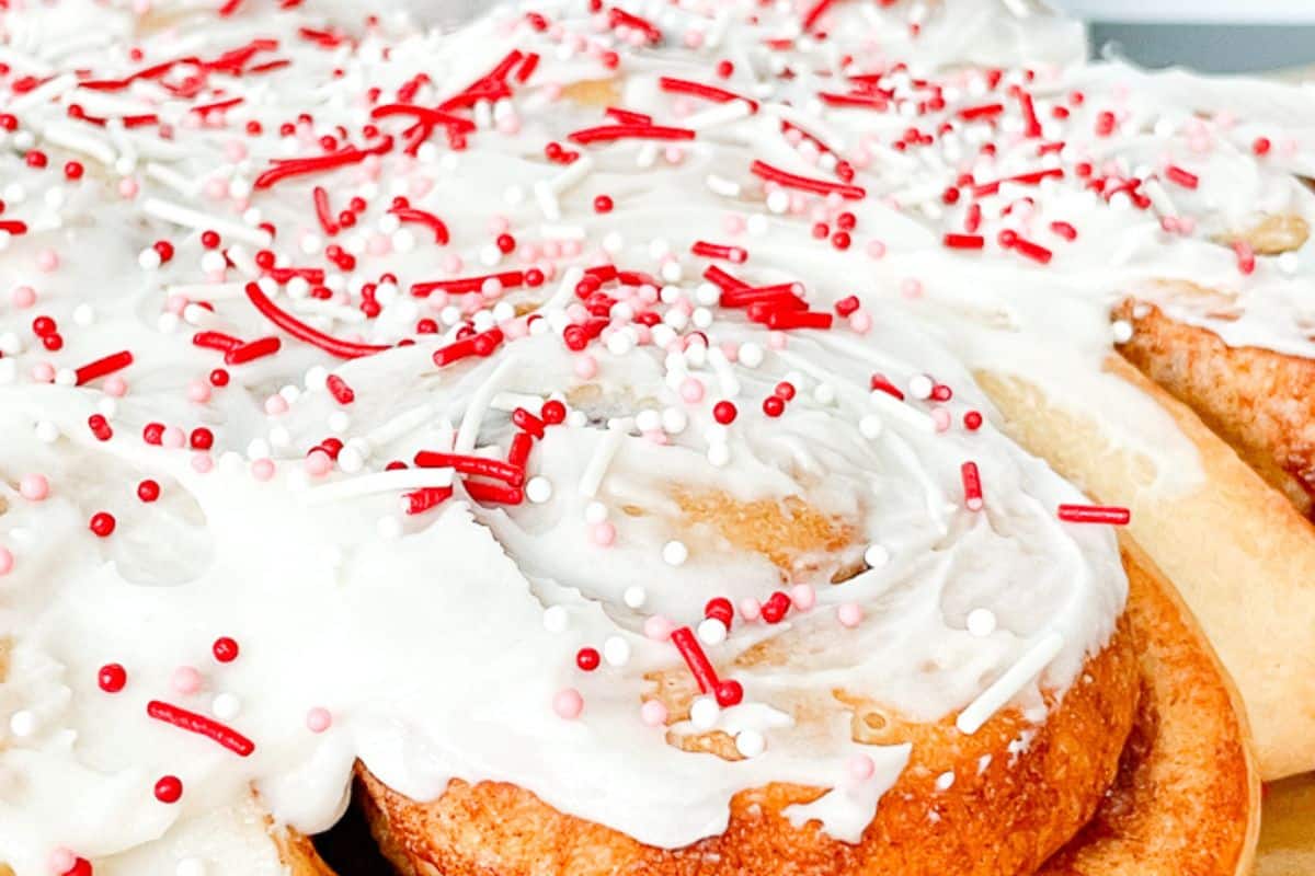 A batch of cinnamon rolls covered in white icing and topped with red sprinkles on a baking sheet.