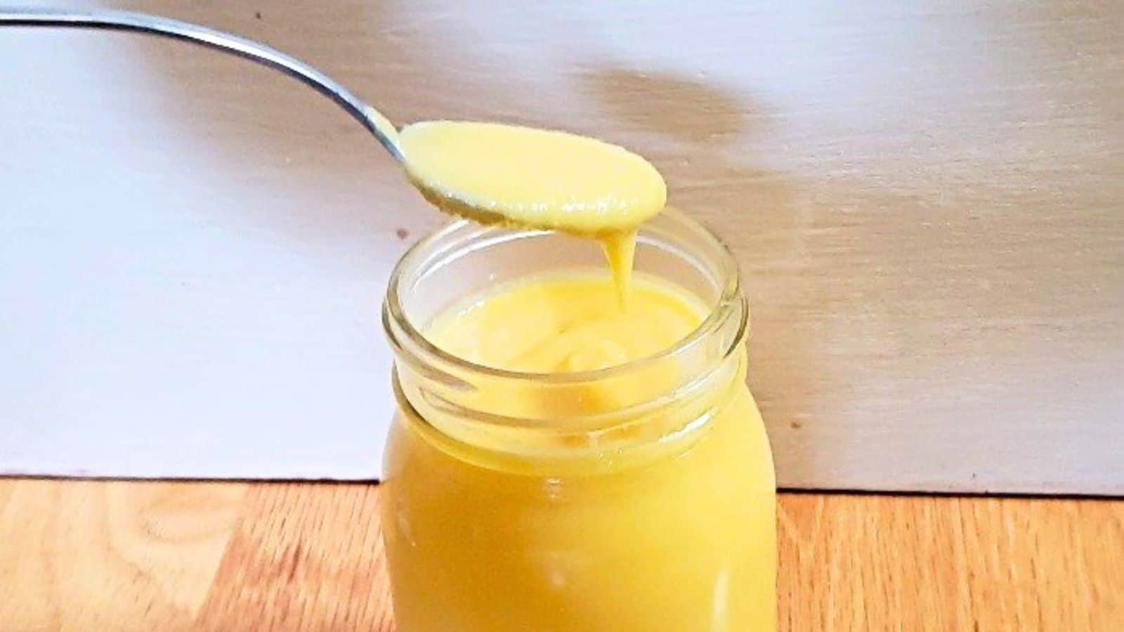 A front on photo shows mango curd in a glass jar with a spoon that has removed some curd.