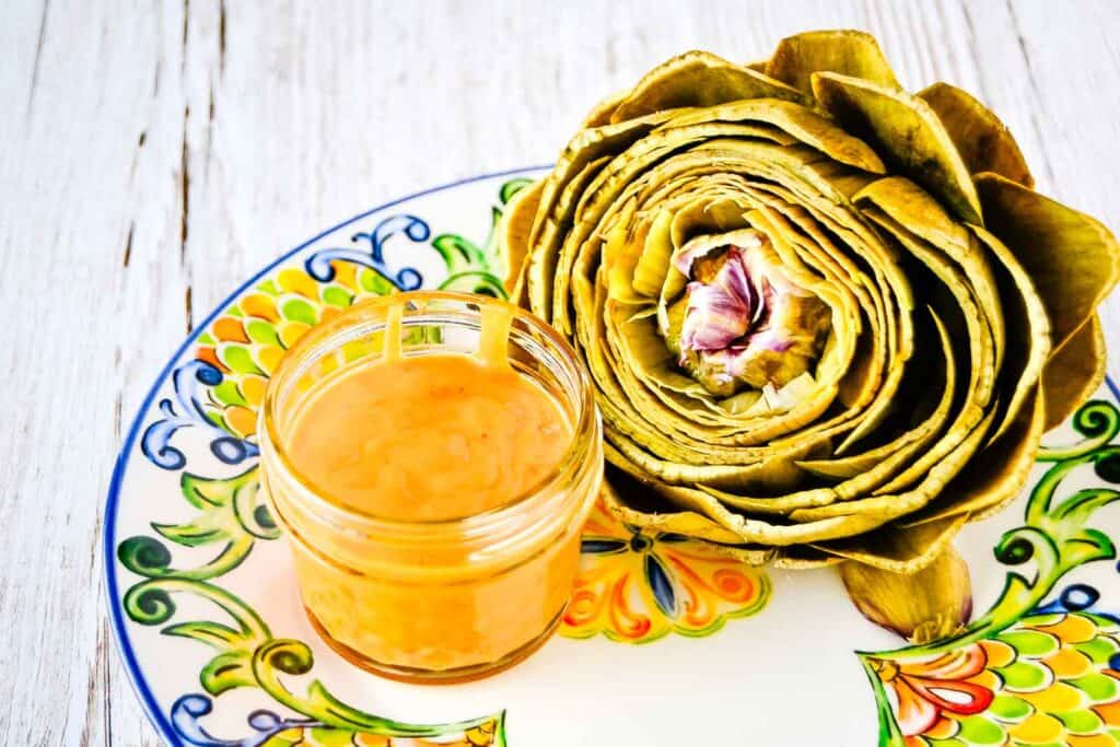 A steamed artichoke on a colorful plate next to a jar of spicy mango sauce.